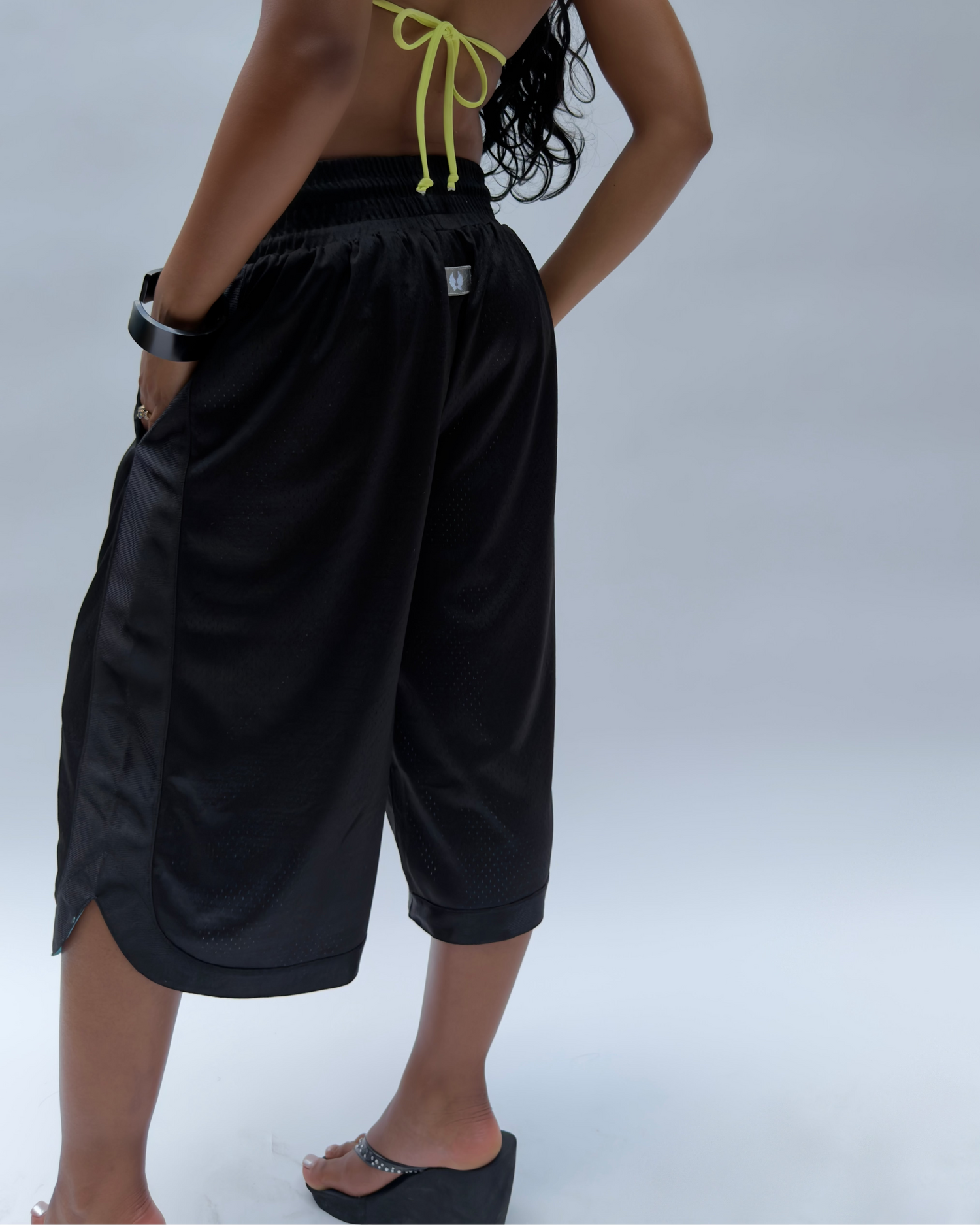 A model shown from the back wearing black wide-leg athletic shorts with side pockets and a neon yellow tie detail on the back, paired with a matching black strapless top and black wedge sandals, against a light gray studio background.