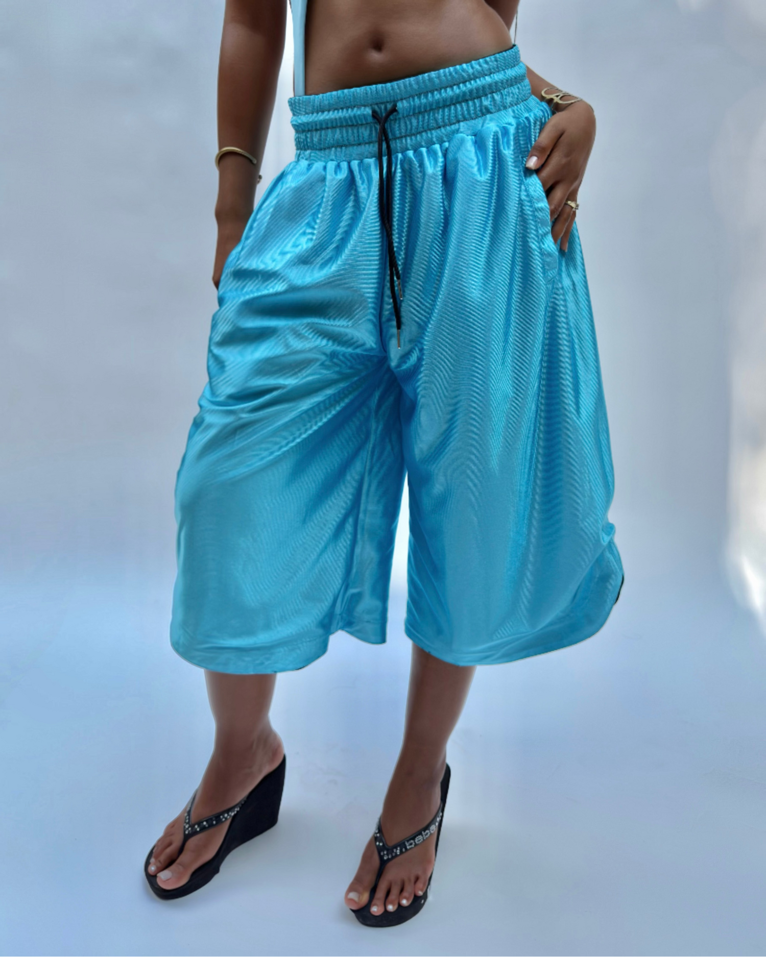 A model wearing bright turquoise blue wide-leg cropped pants with a black drawstring, paired with a matching light blue bikini top and black wedge sandals, set against a light blue studio background.