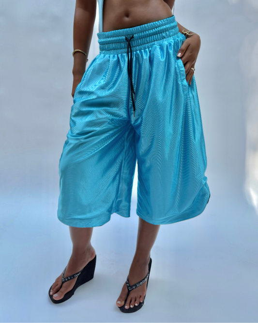 A model wearing bright turquoise blue wide-leg cropped pants with a black drawstring, paired with a matching light blue bikini top and black wedge sandals, set against a light blue studio background.