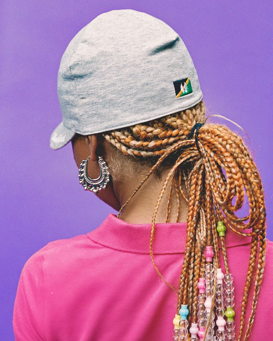 A person shown from the back wearing a light gray cap with a small patch, a bright pink polo shirt, and long braided hair adorned with colorful beads, against a solid purple background.